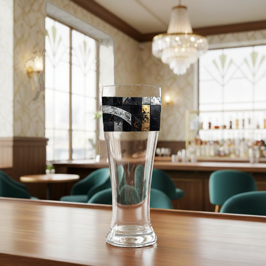 Fresco Fragments pilsner beer glass with a black, silver and gold design on a bar counter with a blurred bar background