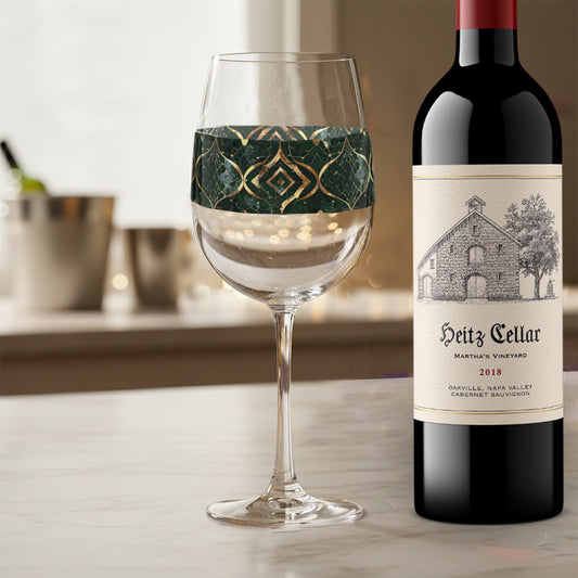 Malachite Enigma tall wine glass with dark green and gold design on a marble counter next to a bottle of Heitz Cellar red wine on a table.