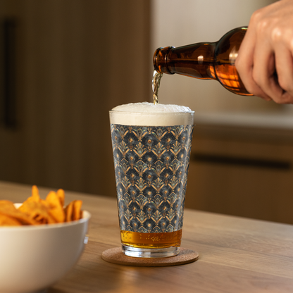 Astral Glow pint beer glass with  a black and bronze design, and a person pouring beer next to a bowl of chips on a wooden table.
