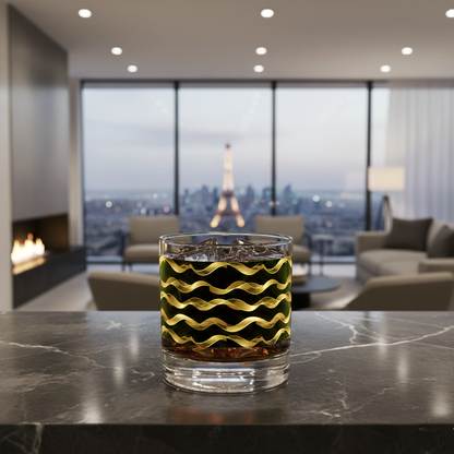 Liquid Gold whiskey glass with gold and black wavy design on a dark marble counter in a modern living room with large city view, fireplace and sectional