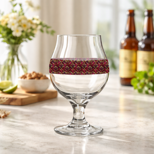 Crimson Jewels Belgian beer glass with a ruby and gold design on a marble surface with blurred bottles in the background
