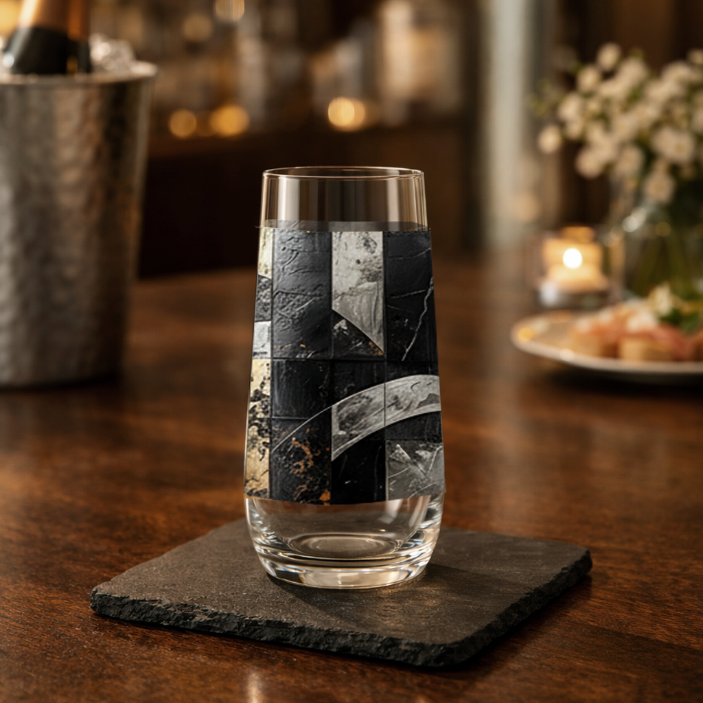 Fresco Fragments stemless champagne glass with black, silver and gold pattern on wood table with a champagne bucket and a blurred home bar in background
