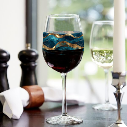 Opulent Teal wine glass with teal and gold design and filled with red wine on a table with a place settings and a blurred curtain and the ouroods in the background