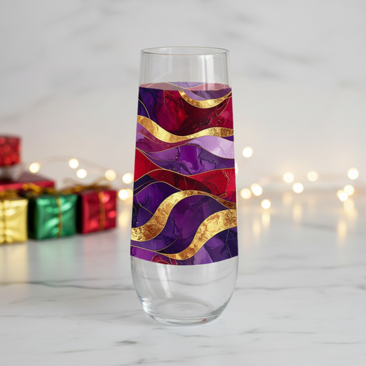 Fluid Marble stemless champagne glass with purple and gold design on a marble surface with blurred holiday gift boxes and lights in the background.