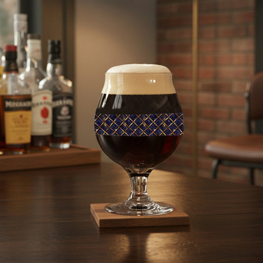 Regal Twilight Belgian beer glass with sapphire and gold wrap designG and filled with dark beer on a coaster, surrounded by bottles in a bar setting.