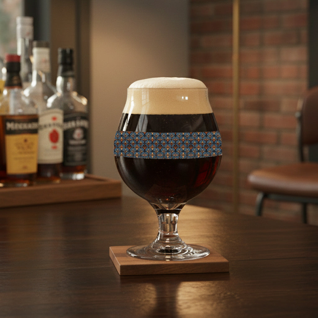 Burnished Slate Belgian beer glass with blue slate and copper design, and filled with dark beer that sits on wooden coaster, with bottles in the background.
