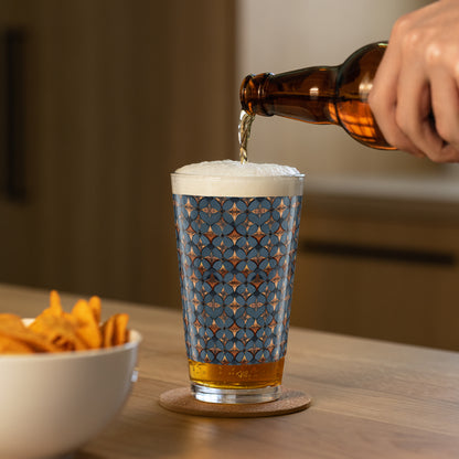 Burnished Slate pint beer glass with slate blue and copper design and a person pouring beer alongside a bowl of chips on a wooden table.